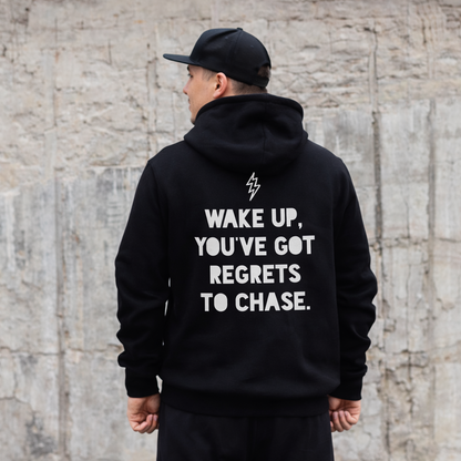 Person wearing a black hoodie with a motivational quote on the back, standing against a textured wall.